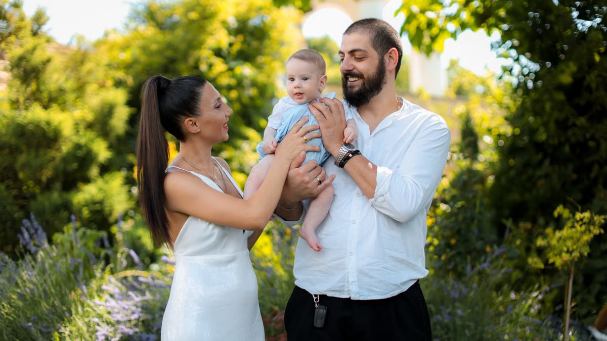 Mother and father joyfully hold their baby in a vibrant outdoor garden setting.