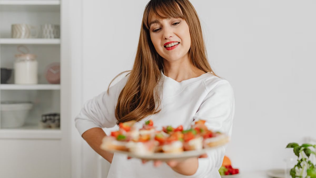 Smiling woman in a white top holding a plate of fresh canapés in a modern kitchen setting.